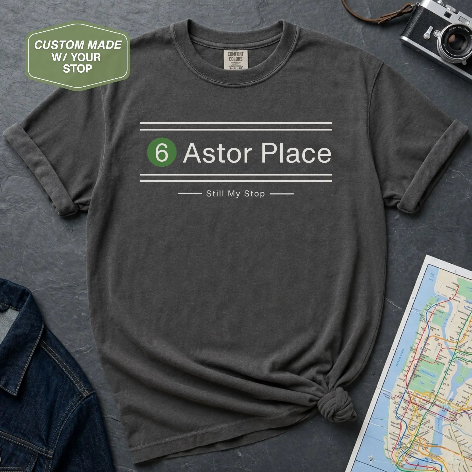 Dark gray t-shirt with '6 Astor Place Still My Stop' text on a textured surface with a map and camera.