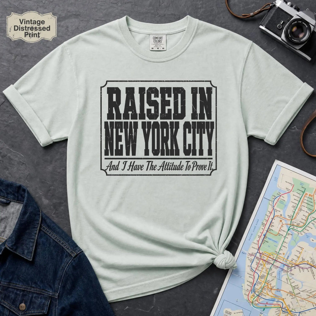 Bay colored t-shirt with 'Raised in New York City' text on a dark surface with a jean jacket and a map.