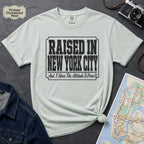 Bay colored t-shirt with 'Raised in New York City' text on a dark surface with a jean jacket and a map.