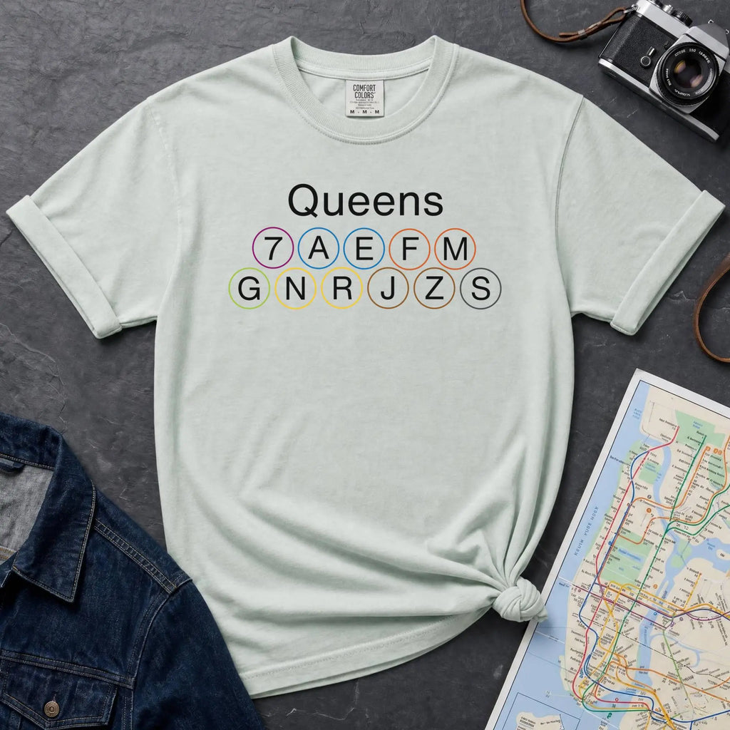 Bay colored t-shirt with 'Queens' and subway line numbers on a dark surface with a map and camera.