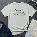 Bay colored t-shirt with 'Queens' and subway line numbers on a dark surface with a map and camera.