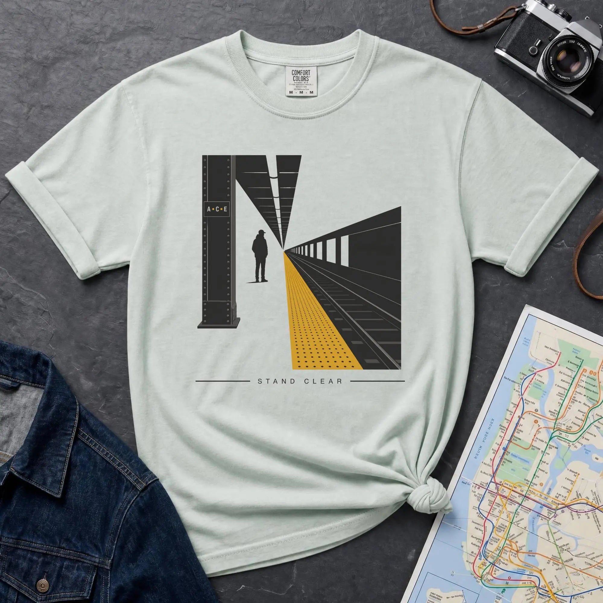 Bay colored t-shirt with a graphic design of a train station platform, denim jacket, and map on a dark surface