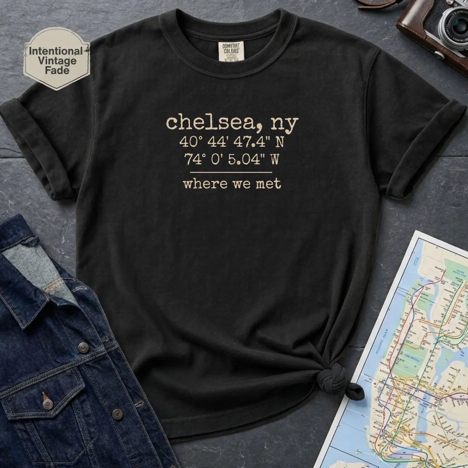 Black Comfort Colors t-shirt with coordinates and text 'chelsea, ny' and 'where we met' on a dark surface with denim jacket and map.