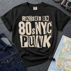Black Comfort Colors t-shirt with bold distressed ransom note style lettering reading "Raised on 80s NYC Punk" in mixed fonts and sizes, black ink on ivory, xerox zine aesthetic.
