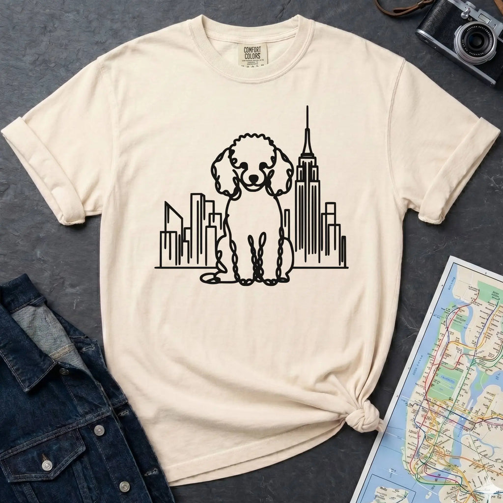 Ivory t-shirt with a poodle and cityscape design on a dark surface with a map and camera.