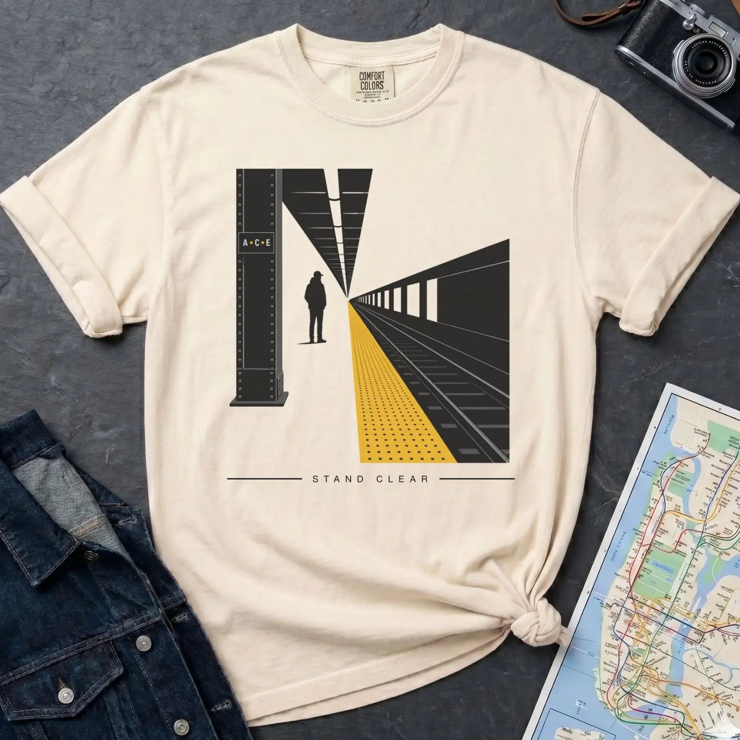 Ivory Comfort Colors t-shirt with a graphic design of a train station platform and text 'Stand Clear' on a dark surface with a denim jacket and a map.