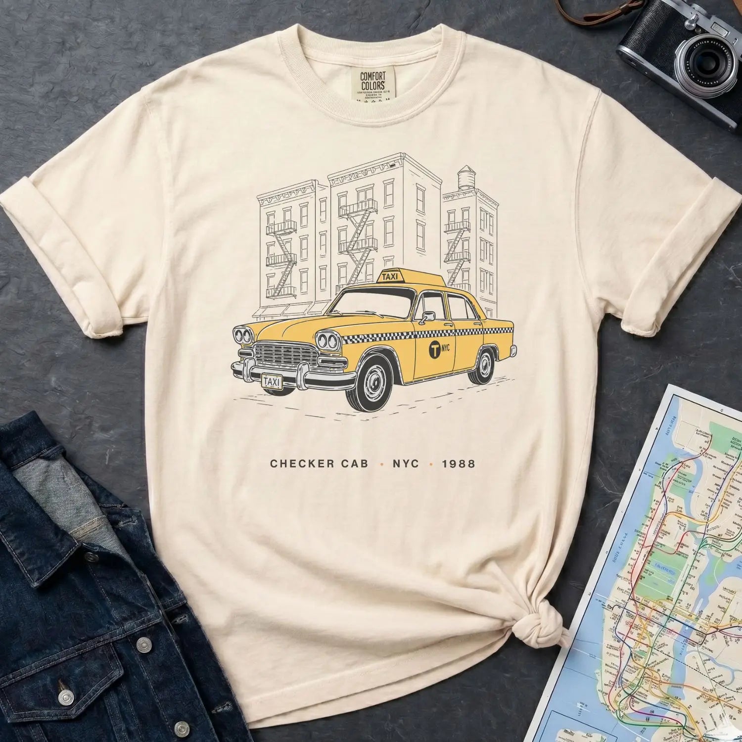 Ivory Comfort Colors t-shirt featuring a vintage New York City Checker Cab graphic with NYC apartment buildings and “Checker Cab • NYC • 1988” text, placed on a dark surface with denim jacket and map.