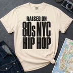 Ivory Comfort Colors T-shirt with black Raised on 80s NYC Hip Hop front print, styled with denim jacket, camera, and subway map
