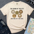 Ivory Comfort Colors t-shirt with 'The Sound of the City' design featuring vintage coins on a textured surface with a camera and map.