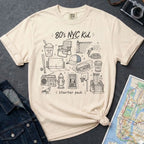 Ivory Comfort Colors t-shirt with '80's NYC Kid' graphic design on a dark surface with denim jacket and map.