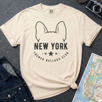 Ivory Comfort Colors t-shirt featuring a New York French Bulldog Club graphic with bold text, outlined French bulldog ears, stars, and a minimalist black design on a gray surface with a map and denim jacket.