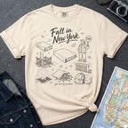 Ivory Comfort Colors t-shirt with 'Fall in New York' graphic design on a dark surface with a denim jacket and a map.