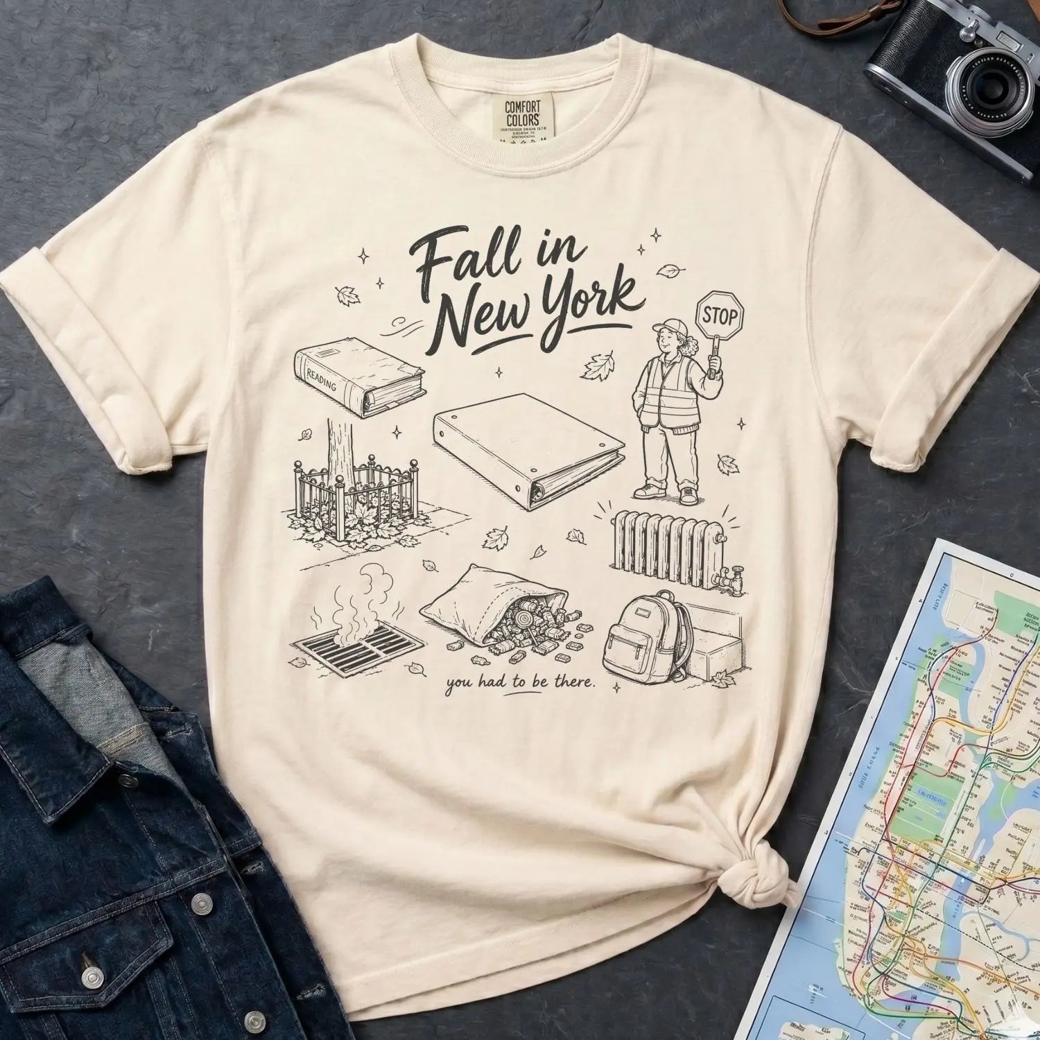 Ivory Comfort Colors t-shirt with 'Fall in New York' graphic design on a dark surface with a denim jacket and a map.
