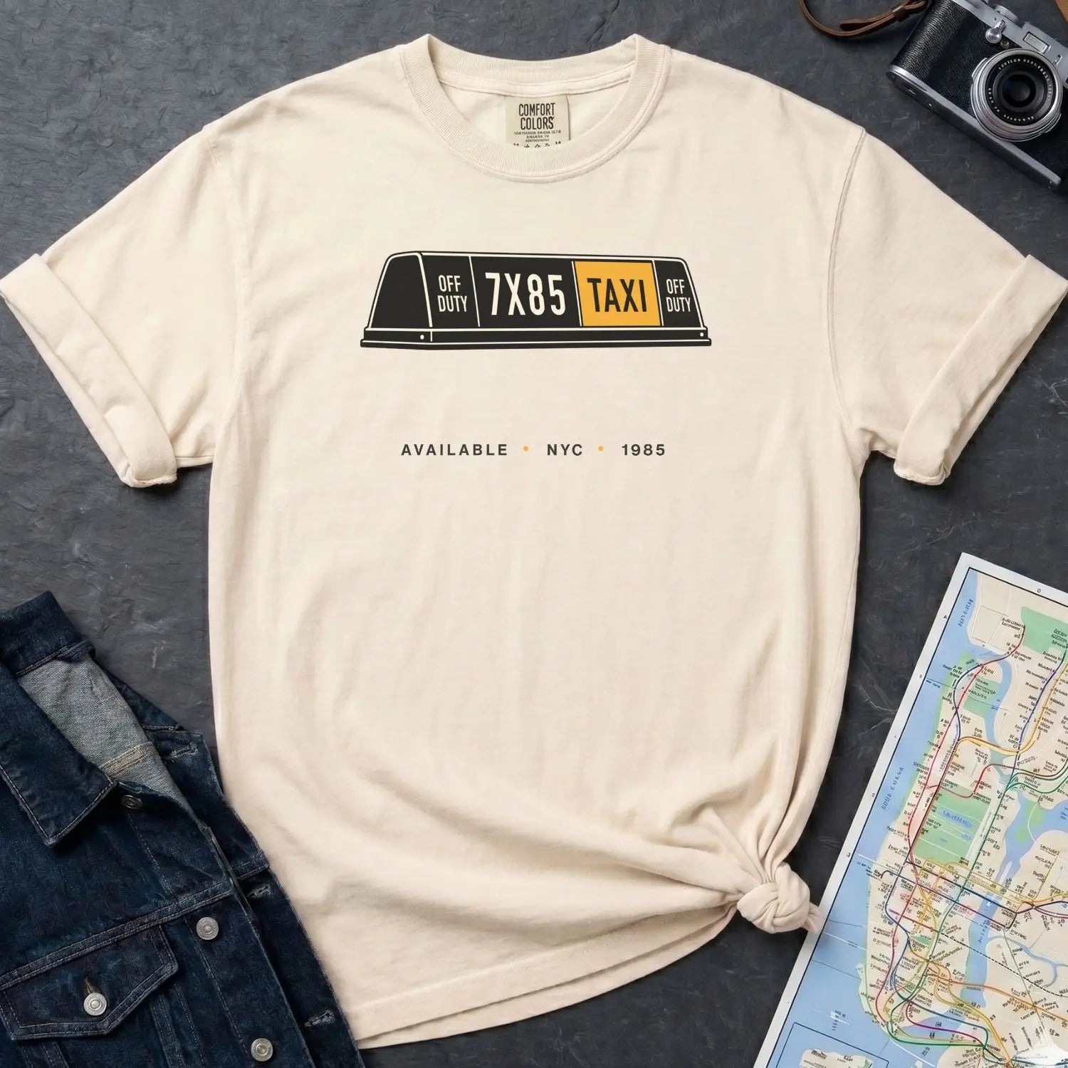 Ivory colored Comfort Colors t-shirt with NYC taxi-themed graphic on a gray surface with a denim jacket and a map.