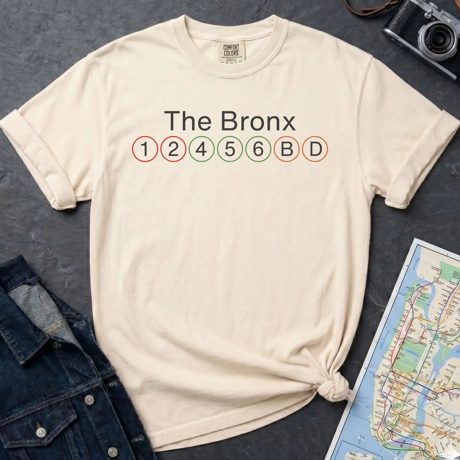 Ivory Comfort Colors t-shirt with 'The Bronx' and subway line text, next to a denim jacket and a map on a dark surface