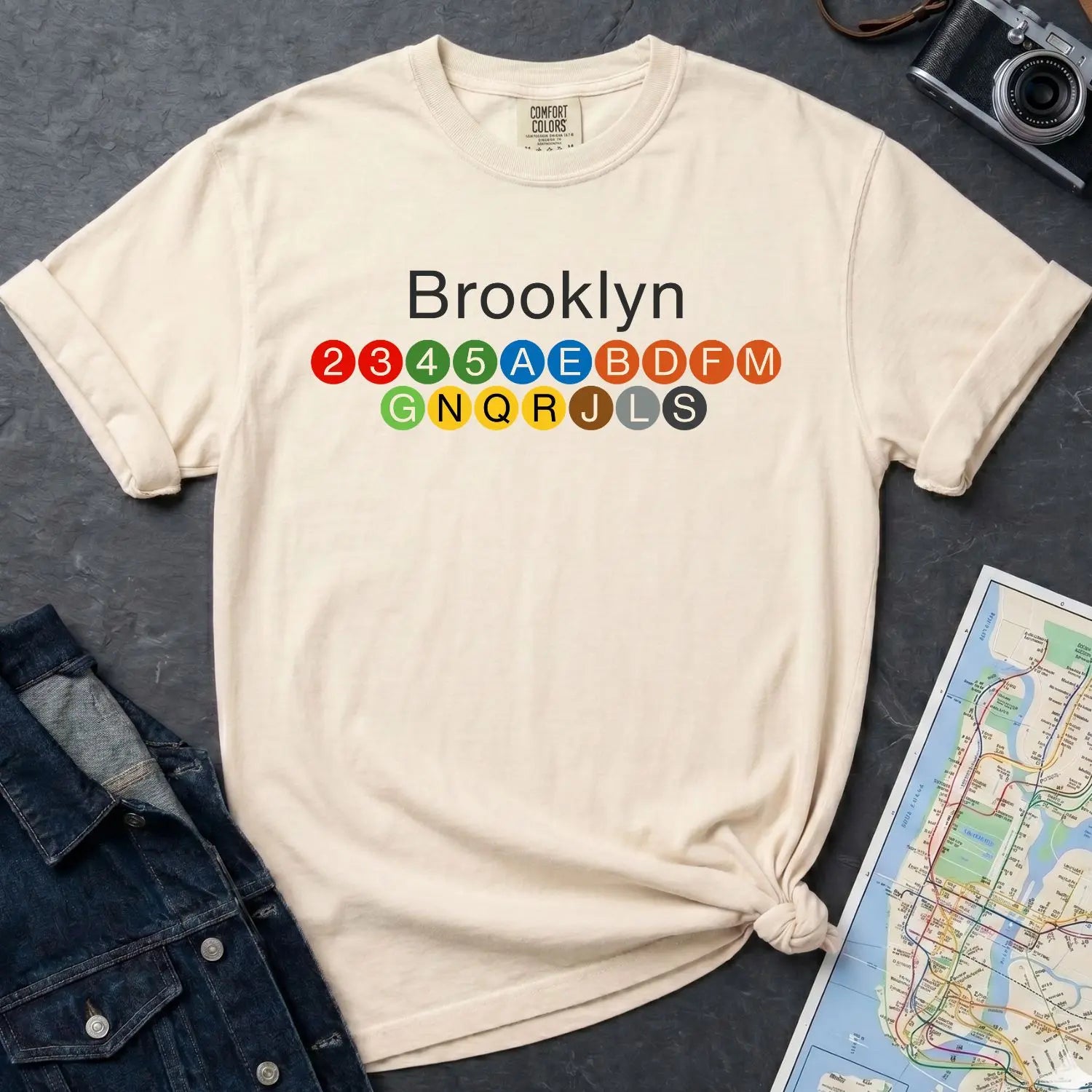Ivory Comfort Colors t-shirt with 'Brooklyn' and subway lines on a dark surface with a map and camera.