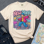 Ivory Comfort Colors t-shirt featuring a colorful NYC graffiti-style New York graphic with a young street artist holding a spray paint can design on a gray surface with denim jacket and map.