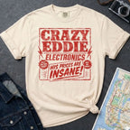 Ivory Comfort Colors t-shirt with distressed vintage retail graphic reading Crazy Eddie's Electronics in bold blocky lettering with lightning bolts and the tagline His Prices Are Insane, styled after 1980s NYC consumer electronics store signage.