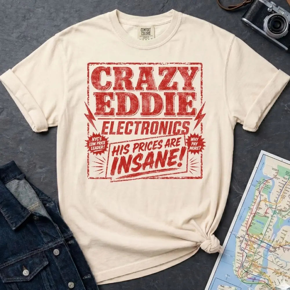 Ivory Comfort Colors t-shirt with distressed vintage retail graphic reading Crazy Eddie's Electronics in bold blocky lettering with lightning bolts and the tagline His Prices Are Insane, styled after 1980s NYC consumer electronics store signage.