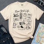 Ivory Comfort Colors t-shirt featuring a New York Life Was the Best Life graphic with hand-drawn NYC nostalgia icons including a MetroCard, cassette tape, pay phone, coffee cup, newspaper, pizza slice, fire hydrant, Walkman, and bodega cat.