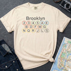 Ivory Comfort Colors t-shirt with Brooklyn subway line design on a dark surface with a denim jacket and a map.