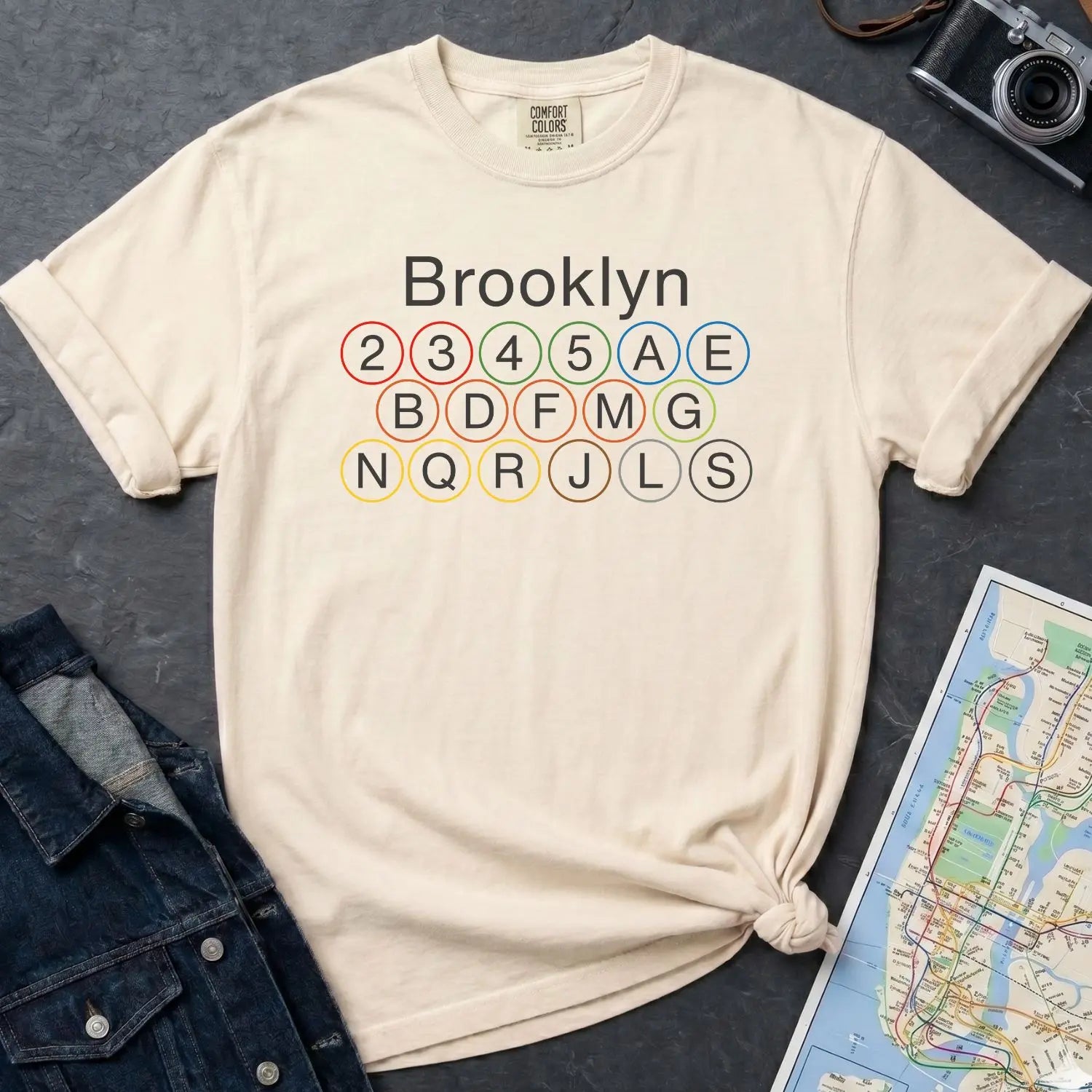 Ivory Comfort Colors t-shirt with Brooklyn subway line design on a dark surface with a denim jacket and a map.