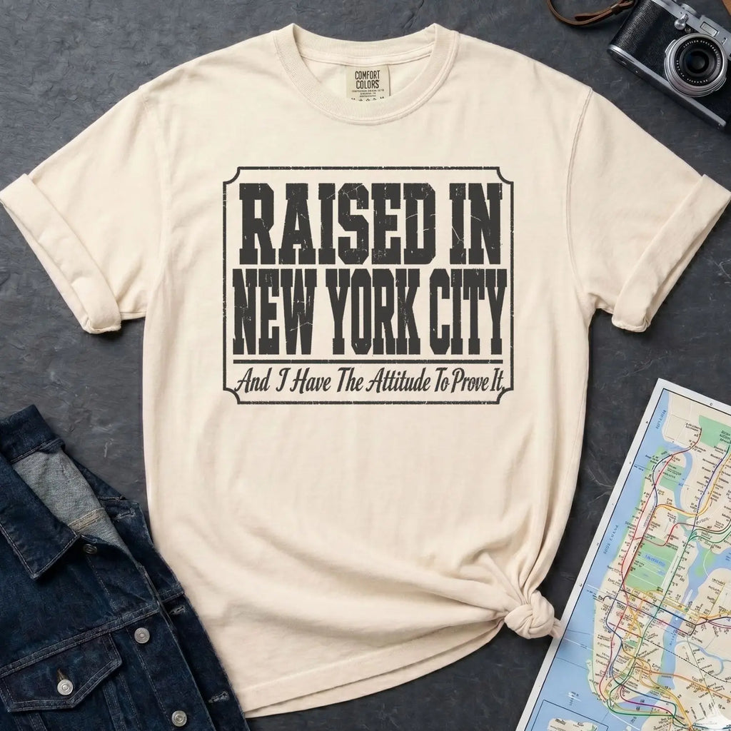 Ivory Comfort Colors t-shirt with 'Raised in New York City' text on a dark surface with a map and denim jacket.