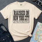 Ivory Comfort Colors t-shirt with 'Raised in New York City' text on a dark surface with a map and denim jacket.
