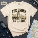 Ivory t-shirt with 'The Bronx Est. 1898' design on a dark surface with a map and denim jacket.