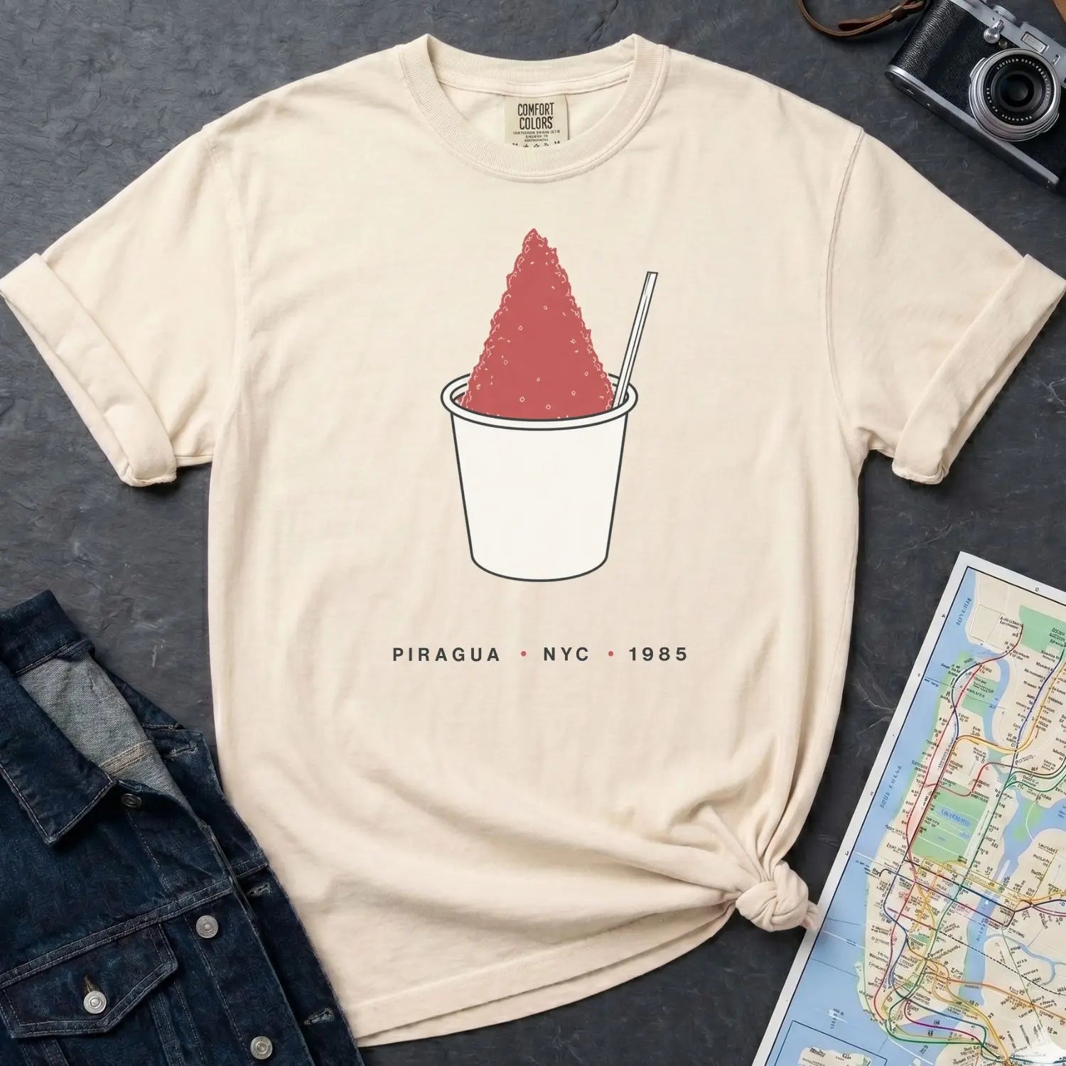 Ivory Comfort Colors t-shirt with a piragua graphic and text, placed on a dark surface with a map and denim jacket.