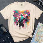 Ivory Comfort Colors t-shirt with a colorful graphic of a person spray painting a brick wall, placed on a dark surface with a map and denim jacket.
