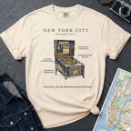 Ivory Comfort Colors t-shirt with pinball machine graphic and text on a dark surface with a denim jacket and map.