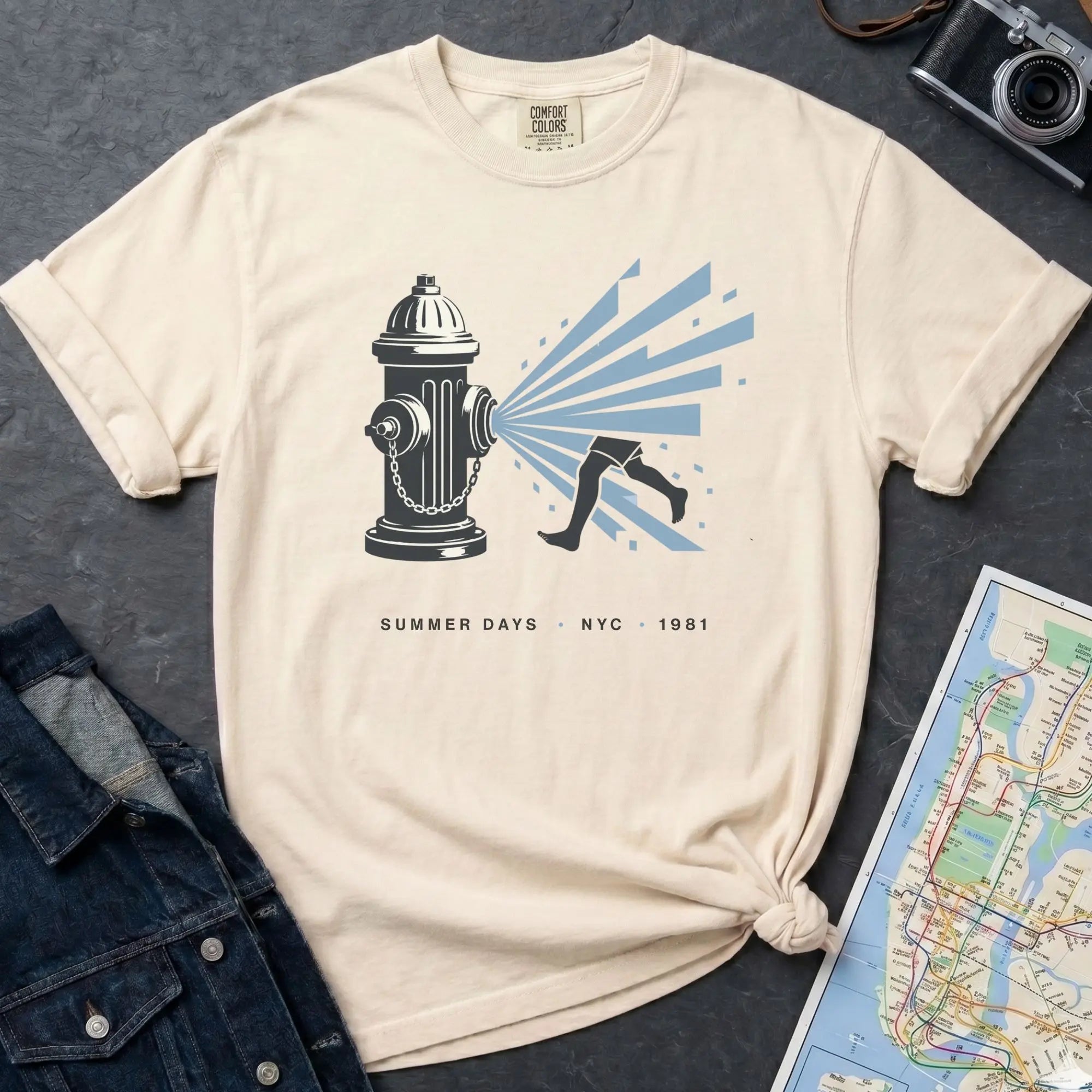 Ivory Comfort Colors t-shirt featuring a vintage NYC fire hydrant spraying water with “Summer Days • NYC • 1981” text, placed on a dark surface with a denim jacket and a map.