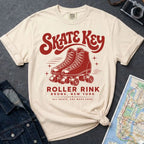 An ivory colored Comfort Colors t-shirt featuring a red retro design that reads 'SKATE KEY' and 'ROLLER RINK BRONX, NEW YORK' with an illustration of roller skates. The shirt is styled in a flat-lay arrangement with a denim jacket, a camera, and a map on a dark gray textured background.