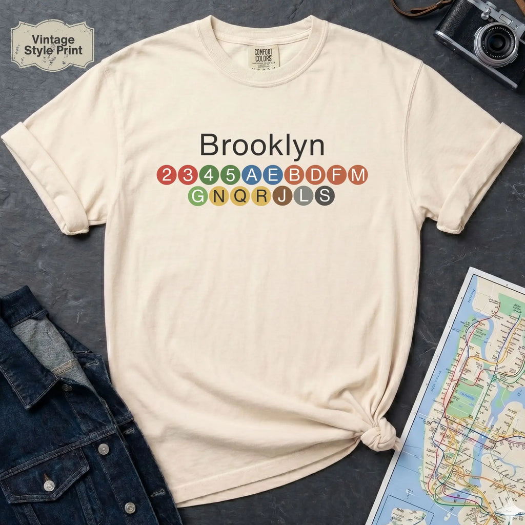 Beige t-shirt with 'Brooklyn' and subway line numbers on a dark surface with a map and camera.