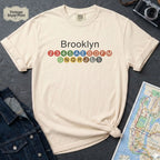 Beige t-shirt with 'Brooklyn' and subway line numbers on a dark surface with a map and camera.