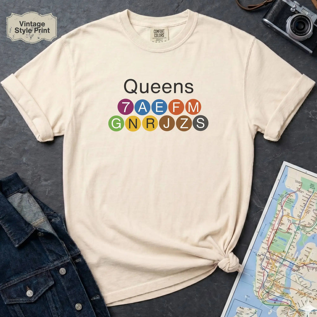 Beige t-shirt with 'Queens' and NYC subway lines on a dark surface with a map and denim jacket.