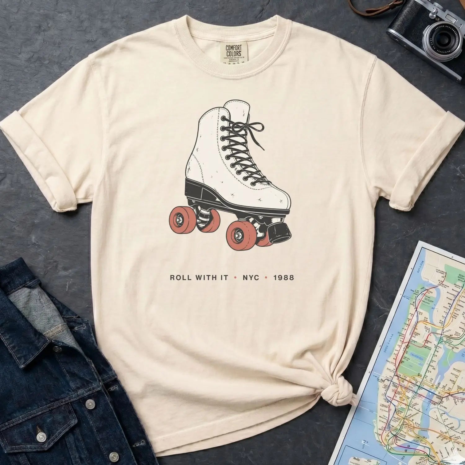Ivory Comfort Colors t-shirt featuring a vintage roller skate graphic with “Roll With It • NYC • 1988” text on a dark surface with a denim jacket and map.