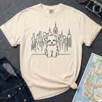 Ivory Comfort Colors t-shirt with a Maltese dog and cityscape design on a gray surface with a map and camera.
