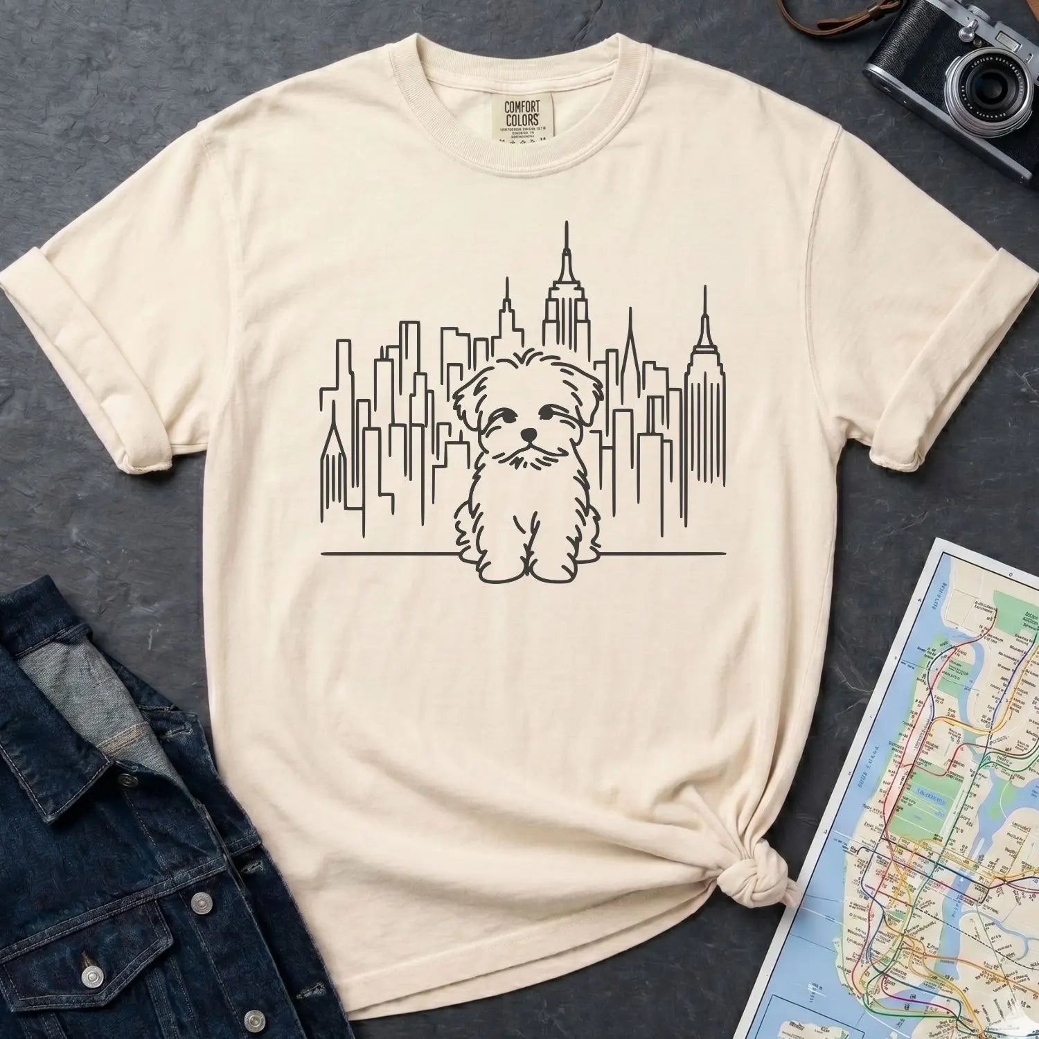 Ivory Comfort Colors t-shirt with a Maltese dog and cityscape design on a gray surface with a map and camera.