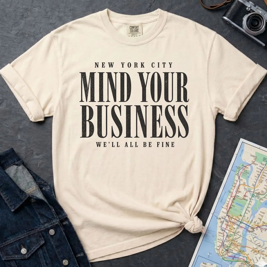 Ivory Comfort Colors t-shirt featuring bold New York City “Mind Your Business, We’ll All Be Fine” text in a vintage black typography design on a dark surface with a denim jacket and a map.