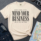 Ivory Comfort Colors t-shirt featuring bold New York City “Mind Your Business, We’ll All Be Fine” text in a vintage black typography design on a dark surface with a denim jacket and a map.