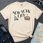 Ivory Comfort Colors t-shirt with 'New York City' text and a roller skate and boom box on a dark surface with a denim jacket and a map.