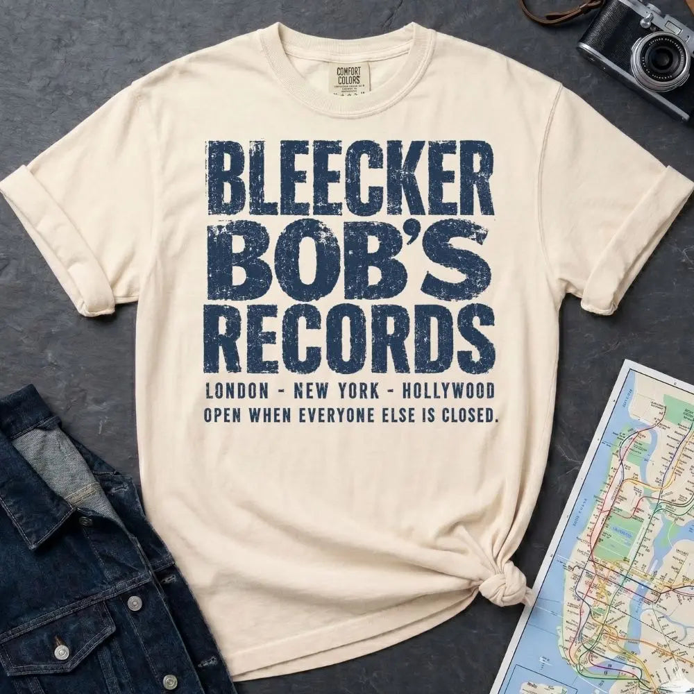 Ivory Comfort Colors t-shirt with Bleecker Bob's Records in large distressed block lettering, London, New York, Hollywood and Open When Everyone Else Is Closed below, displayed on a flat surface with a denim jacket and subway map in the background.