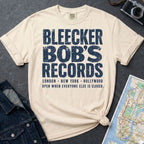 Ivory Comfort Colors t-shirt with Bleecker Bob's Records in large distressed block lettering, London, New York, Hollywood and Open When Everyone Else Is Closed below, displayed on a flat surface with a denim jacket and subway map in the background.