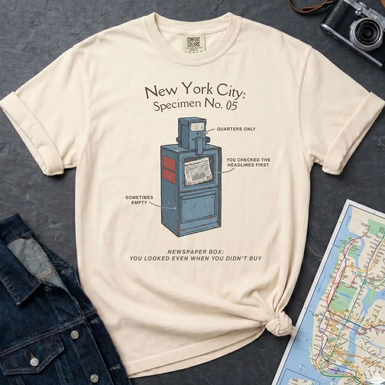 Ivory Comfort Colors t-shirt with a newspaper box graphic and text on a dark surface with a denim jacket and a map.