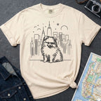Ivory Comfort Colors t-shirt featuring a fluffy Pomeranian dog illustration in front of the New York City skyline on a dark surface with denim jacket and map.