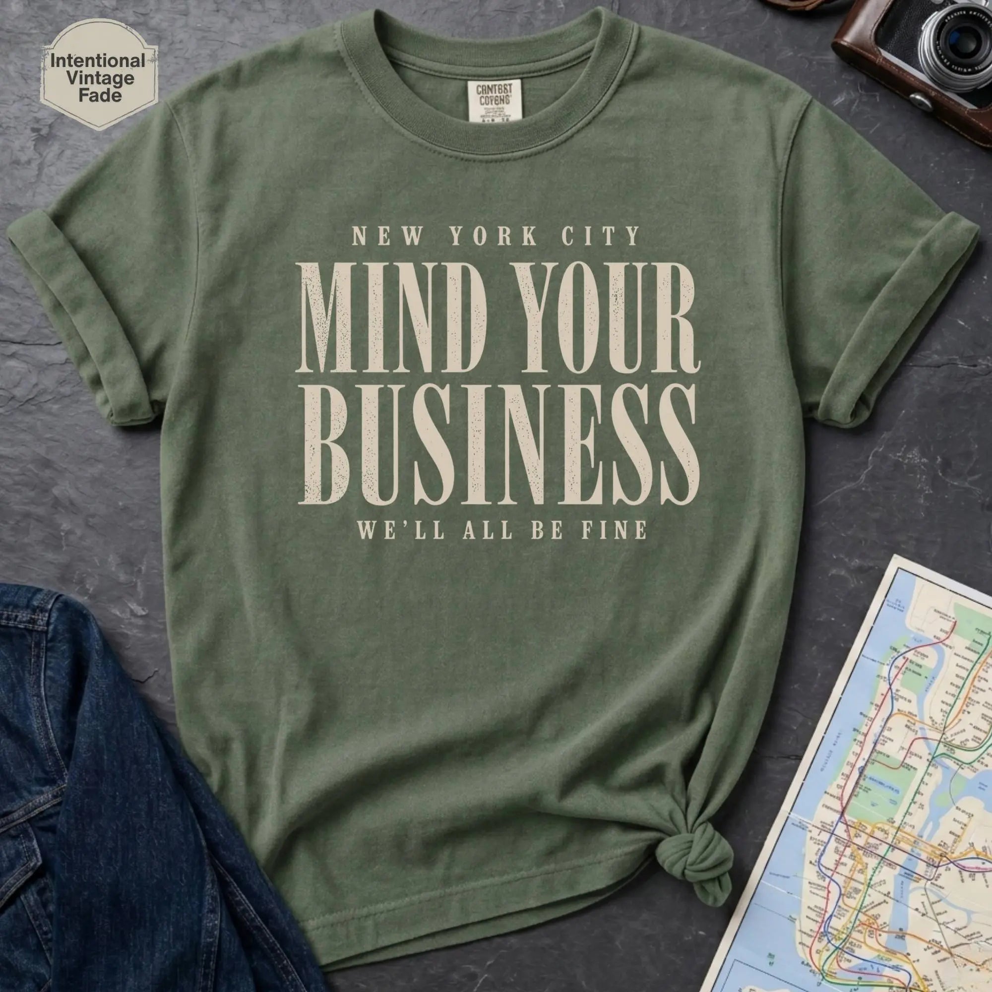 Moss green t-shirt with 'New York City Mind Your Business We'll All Be Fine' text on a dark surface with a map and camera.
