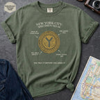 Moss green t-shirt with New York City token design on a gray surface with a map and camera.