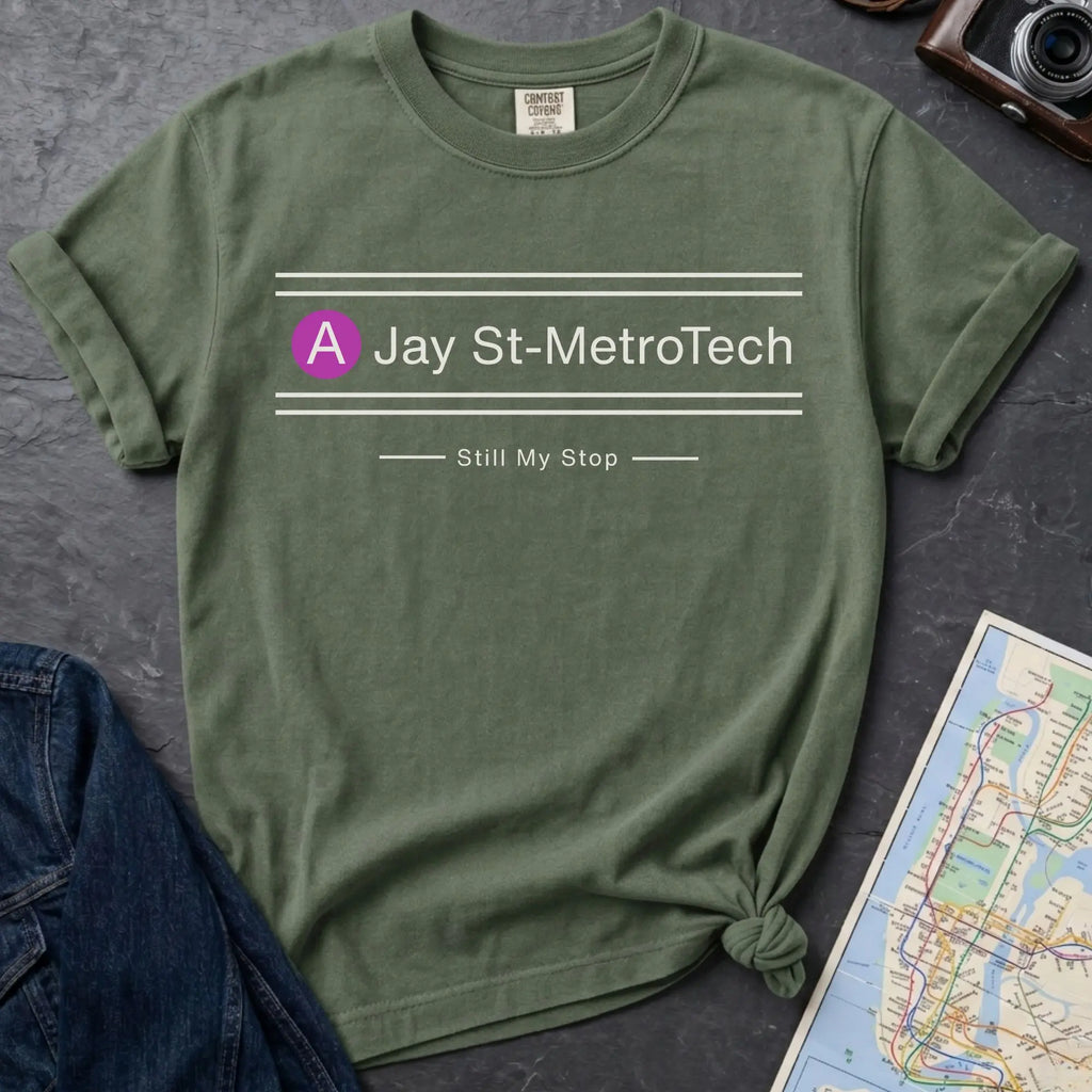 Green t-shirt with 'Jay St-MetroTech Still My Stop' text on a dark surface with a map and jeans.